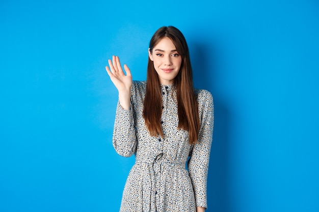 Linda mulher sorridente, acenando com a mão para cumprimentá-lo, dizendo Olá com um sorriso tímido, em um vestido contra o azul.