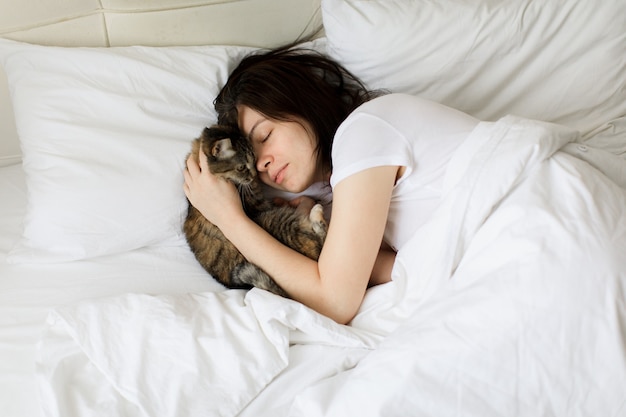 Linda mulher sorridente com os olhos fechados, deitada na cama e abraçando o gato