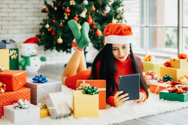 Linda mulher sorridente e feliz no vestido vermelho e com um chapéu de Papai Noel está deitada no chão, cercada por caixas de presente coloridas com uma árvore de Natal no fundo, segurando o tablet na mão.