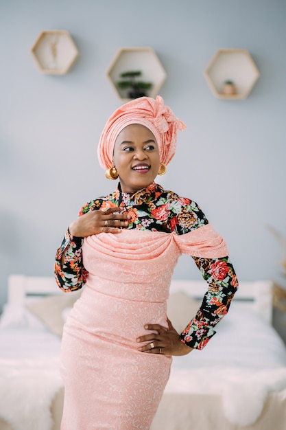 Linda mulher sorridente emocional com maquiagem brilhante contra o fundo claro do estúdio. foto de estúdio da modelo africana feminina em vestido rosa étnico e turbante, olhando para a câmera e sorrindo.