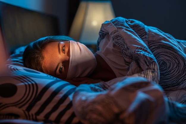 Linda mulher usando uma máscara médica na cama durante o período de quarentena do vírus corona. Falta de liberdade