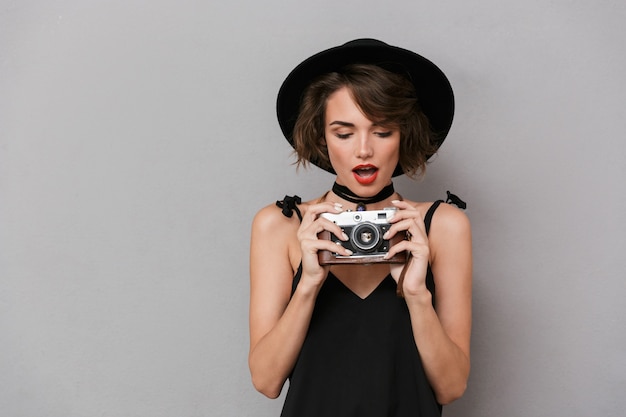 Linda mulher usando vestido preto e chapéu fotografando na câmera retro, isolada sobre uma parede cinza