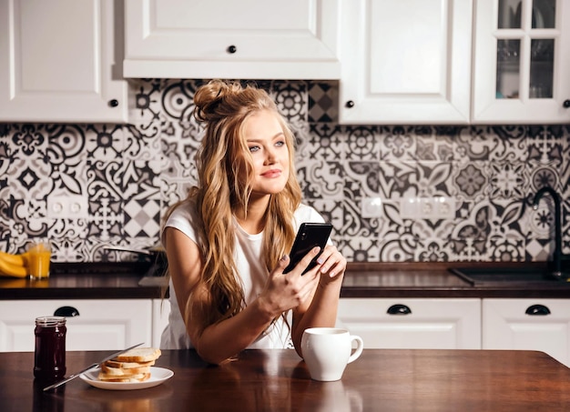 Linda mulher vestindo pijama bebendo chá e usando smartphone na cozinha