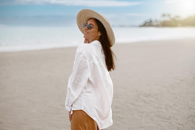 Linda mulher viajante com um chapéu parado na praia tropical e olhando para longe o conceito de férias