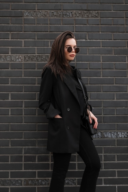 Linda na moda jovem urbana em óculos de sol redondos da moda em uma jaqueta elegante está de pé perto da parede de tijolo preto vintage na cidade. Modelo de moda linda garota moderna em vestido casual ao ar livre.