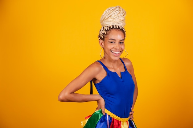 Linda negra brasileira vestida com roupas de carnaval coque de fundo azul e amarelo e tranças com as mãos na cintura