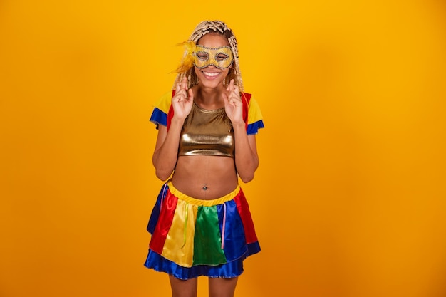 Linda negra brasileira vestida com roupas de carnaval máscara fundo amarelo dedos cruzados desejando esperança