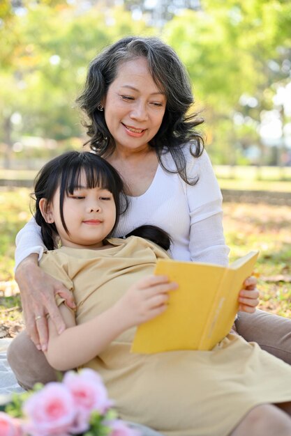Linda nieta asiática acostada sobre su abuela leyendo un libro mientras hace un picnic en el parque