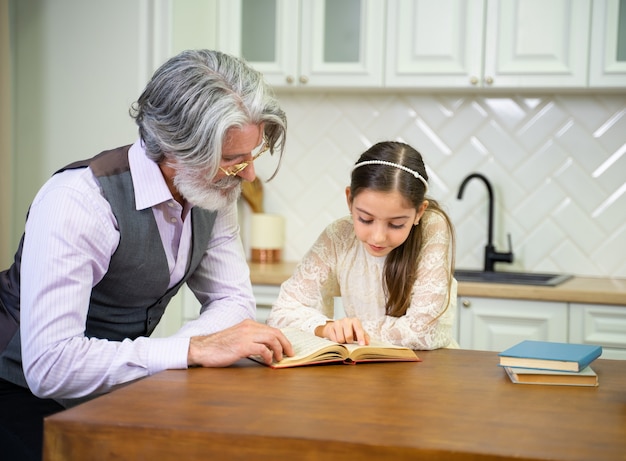 linda nieta leyendo un libro de cuentos con el abuelo mientras está sentado en la cocina