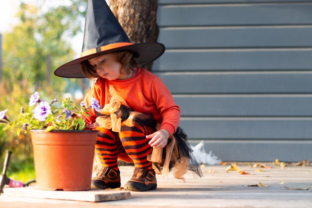 Una linda niña 2-3 en un disfraz de bruja de Halloween naranja y negro se sienta junto a las flores en la terraza de la casa gris