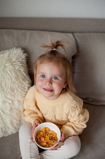 Una linda niña de 2 años en un acogedor suéter de punto está desayunando en el sofá