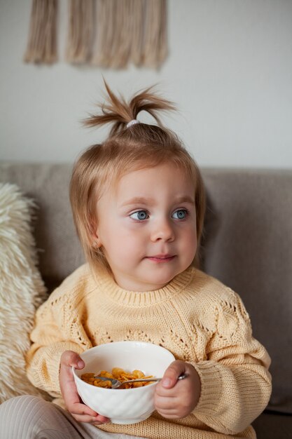 Una linda niña de 2 años en un acogedor suéter de punto está desayunando en el sofá