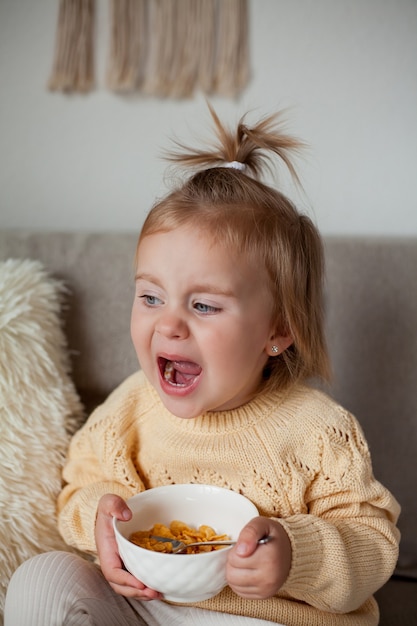 Una linda niña de 2 años en un acogedor suéter de punto está desayunando en el sofá