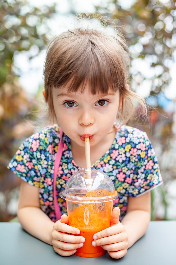 Linda niña de 4 años bebiendo jugo de zanahoria fresco con pajita para sentarse afuera