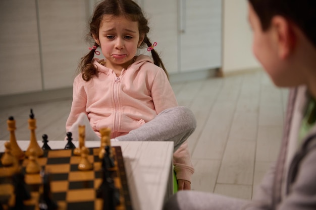 Foto linda niña de 4 años con mirada molesta sentada junto a su hermano y aprendiendo ajedrez