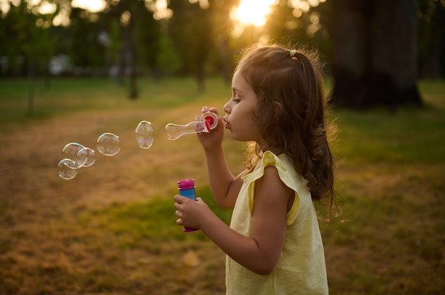 Linda niña de 4 años, soplando pompas de jabón sobre un fondo de parque de la ciudad al atardecer, disfrutando de un rato agradable al aire libre. Los rayos del sol caen a través de esferas de burbujas transparentes con reflejos iridiscentes.