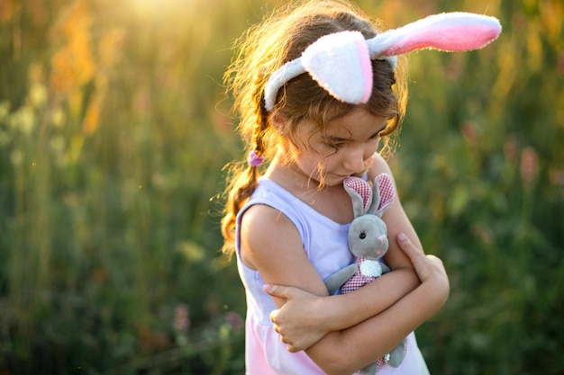 Linda niña de 5 años con orejas de conejo abraza suavemente a un conejo de juguete en la naturaleza en un campo floreciente en verano con luz dorada. Pascua, conejito de Pascua, infancia, niño feliz, primavera.