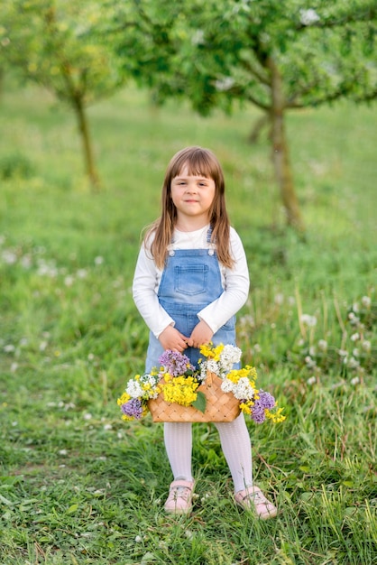 Una linda niña de 5 años con un vestido de mezclilla se para con una canasta de flores en el jardín de primavera