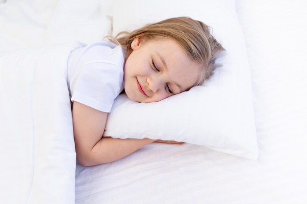 Una linda niña abraza una almohada con las manos dobladas debajo de la mejilla y duerme en la cama en una cama de algodón blanco debajo de una manta, el sueño de un bebé saludable por la noche