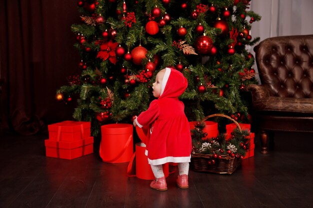 Linda niña de un año en traje de Santa Claus en el fondo del árbol de Navidad decorado con juguetes. Niño en adornos con cajas de regalo. Concepto de hogar acogedor celebración de año nuevo feliz. Copia espacio