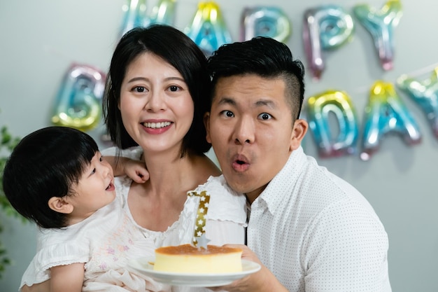 linda niña asiática divirtiéndose mirando a su padre que está soplando velas en el pastel mientras toman una foto familiar con motivo de su primer cumpleaños en casa