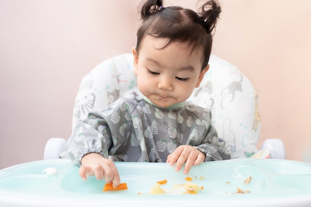 Una linda niña asiática sentada en la mesa de comedor practica comer comida por sí misma, concepto de destete dirigido por bebés