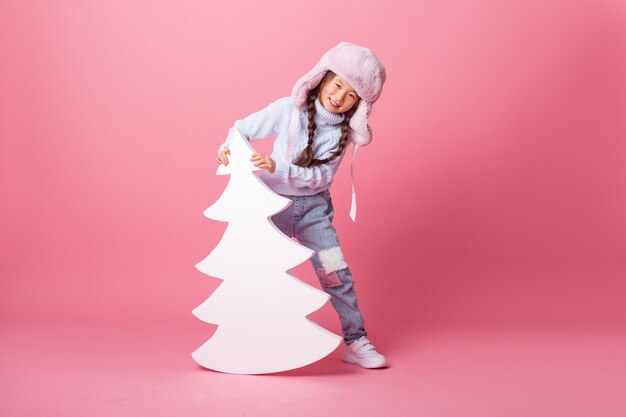 Una linda niña asiática con un sombrero de invierno se encuentra junto a un árbol de Navidad sobre un fondo rosa. concepto de invierno, espacio para texto