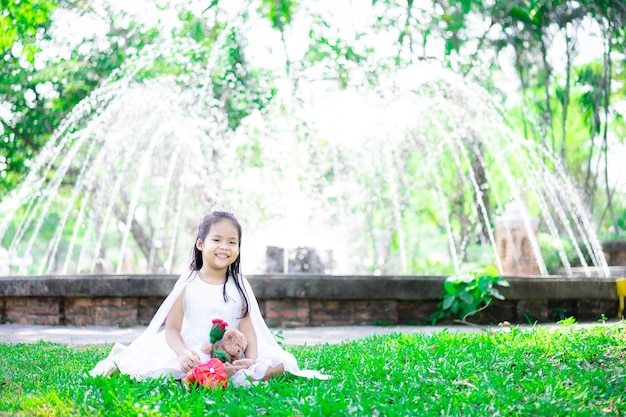 Foto linda niña asiática en vestido blanco con rosa roja y regalo en el parque