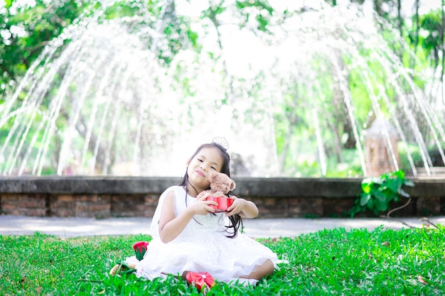 Linda niña asiática en vestido blanco sosteniendo una muñeca de oso en el parque