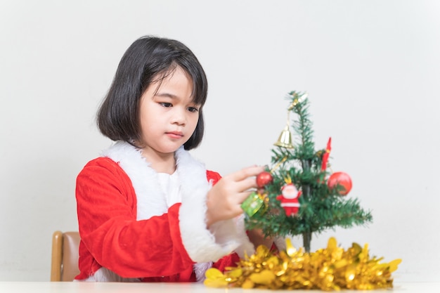 Una linda niña asiática en un vestido rojo de Santa Claus decorando el árbol de Navidad en la víspera de Navidad