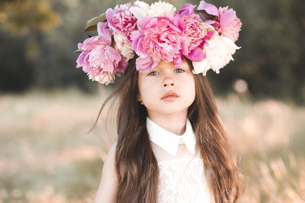 Linda niña bonita con corona floral de flores de peonía posando en el prado al aire libre