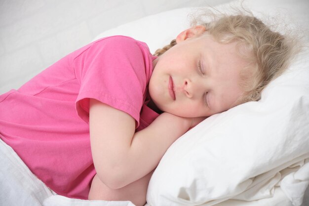 Linda niña con cabello rubio y una camiseta rosa duerme en una cama blanca