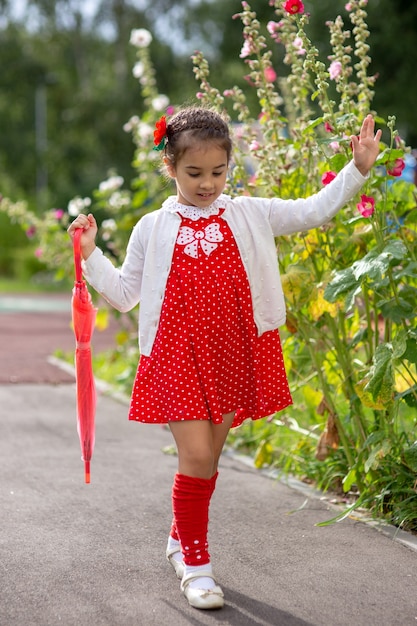 Una linda niña camina con un vestido rojo y una blusa blanca con un paraguas rosa doblado en un día de verano