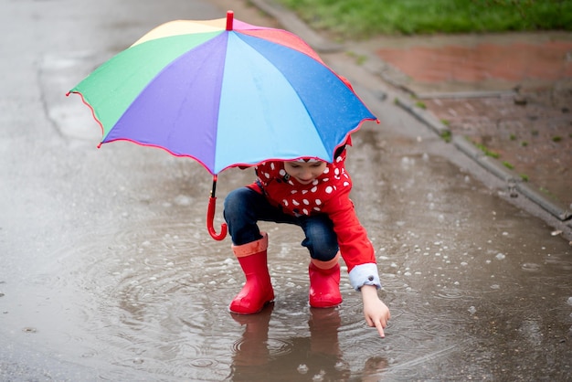 Una linda niña con una capa roja, botas rojas y un sombrero blanco salta en charcos y se divierte