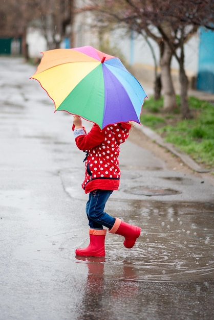 Una linda niña con una capa roja, botas rojas y un sombrero blanco salta en charcos y se divierte