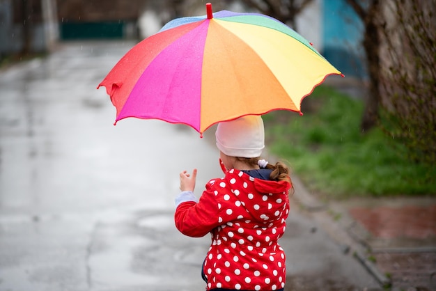 Una linda niña con una capa roja, botas rojas y un sombrero blanco salta en charcos y se divierte