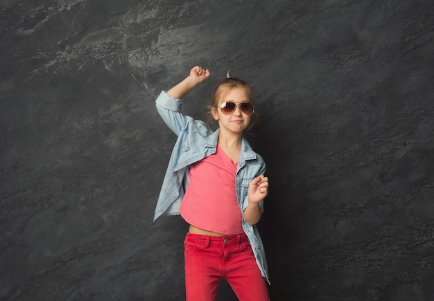 Linda niña casual feliz en gafas de sol divirtiéndose en el fondo negro del estudio, espacio de copia. Expresión de la cara de niña