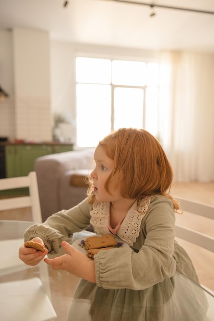 Linda niña caucásica con cabello rojo come galletas caseras mientras se sienta en la mesa Concepto de comida