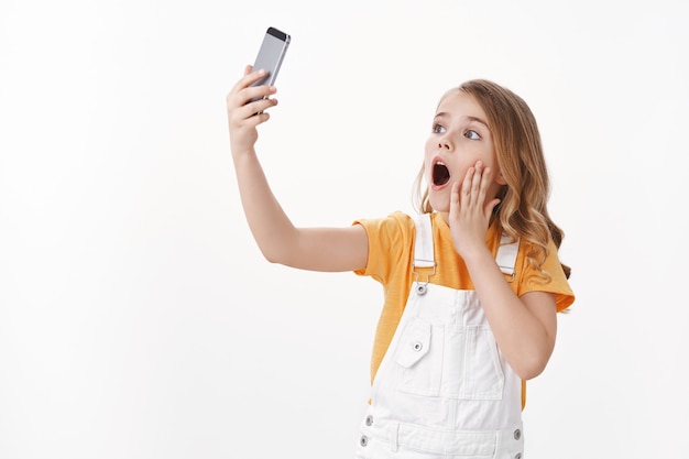 Linda niña caucásica carismática, niño jugando con el teléfono inteligente haciendo muecas divertidas y juguetonas, extender el brazo sujetar el teléfono móvil tomando selfie con emoción sorprendida y asombrada, pared blanca