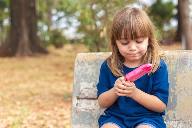 Linda niña caucásica chupando una paleta en el banco del parque al aire libre