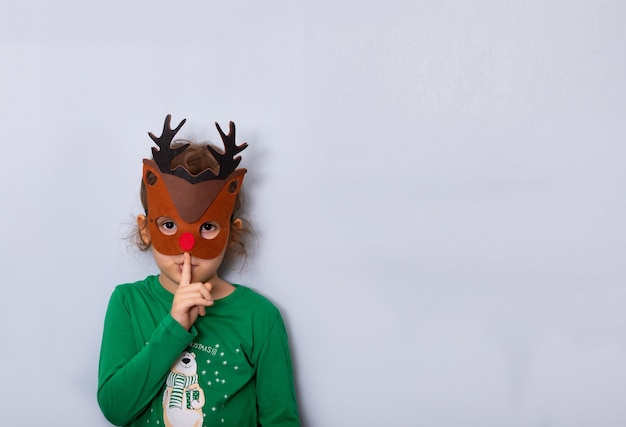 Linda niña caucásica pequeña con máscara de venado haciendo un dedo en los labios gesto de silencio en la boca manteniendo un secreto Copiar espacio para diseño o texto Plantilla de maqueta de año nuevo de banner