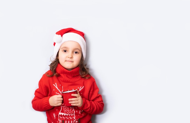 Linda niña caucásica con sombrero rojo de Papá Noel sosteniendo una taza roja o una taza vacía copia espacio para diseño o texto Plantilla de maqueta de Navidad de banner