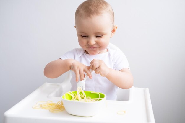 linda niña comiendo pasta de espagueti sentada en una silla de bebé en la cocina blanca