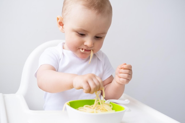 linda niña comiendo pasta de espagueti sentada en una silla de bebé en la cocina blanca