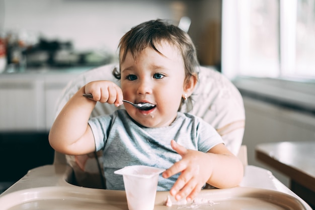 Linda, niña comiendo yogur en la cocina durante el día en una silla alta