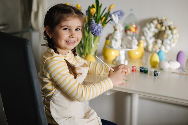 Linda niña de cuatro años en delantal beige sentada en la mesa de la cocina y pintando huevos de Pascua