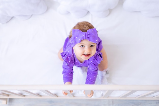 Linda niña en una cuna se aferra a un lado con ropa lila y con un lazo en la cabeza divertido concepto de vista superior de bebé de artículos para niños