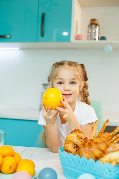 Linda niña divertida juguetona felizmente sosteniendo naranjas naranjas, con cocina en el fondo