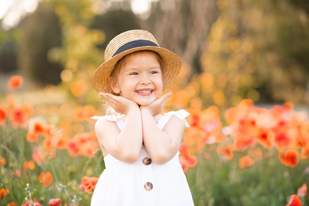 Linda niña divertida usa sombrero de paja y vestido blanco de verano sobre fondo de pradera de flores