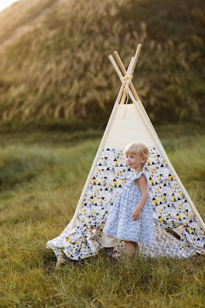 linda niña se divierte cerca de wigwam en un campo de verano al atardecer.